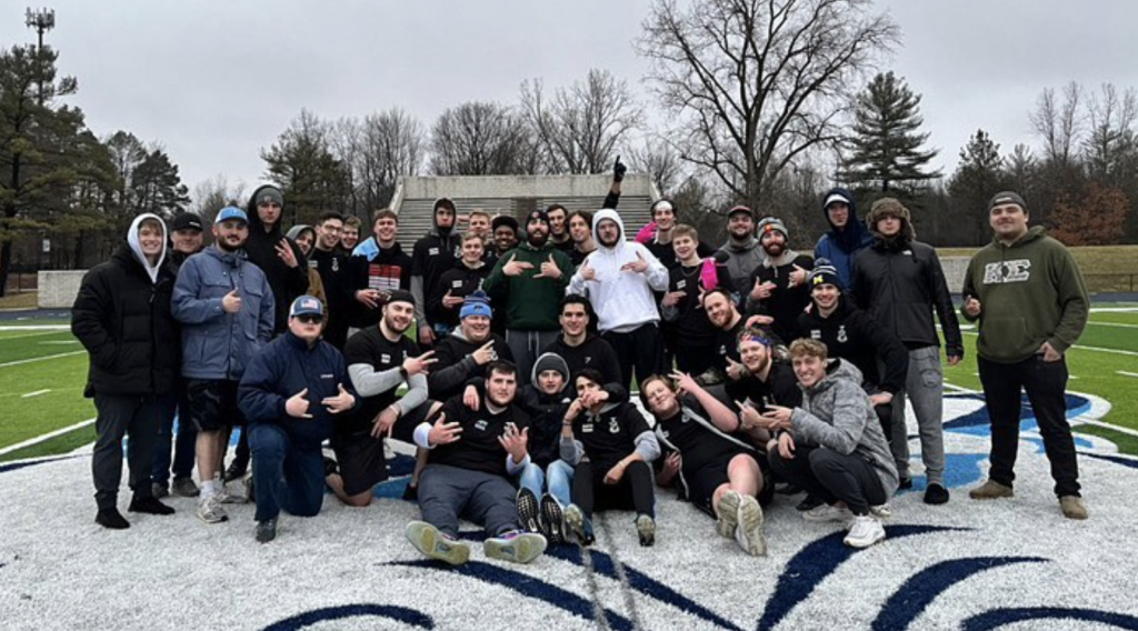 A large group of people, dressed warmly, gathered on a football field, posing and smiling for the camera. Trees without leaves are in the background under an overcast sky, capturing a memorable moment of Greek Life camaraderie.